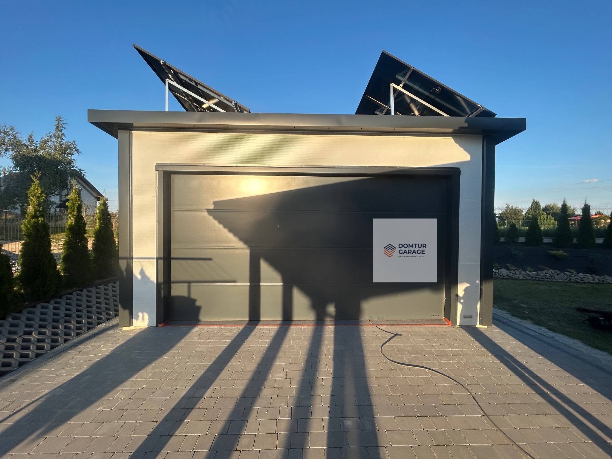 Nowoczesny garaż blaszany z panelami słonecznymi na dachu, oświetlony popołudniowym słońcem, z widocznym logo producenta na bramie. Kostka brukowa przed garażem.