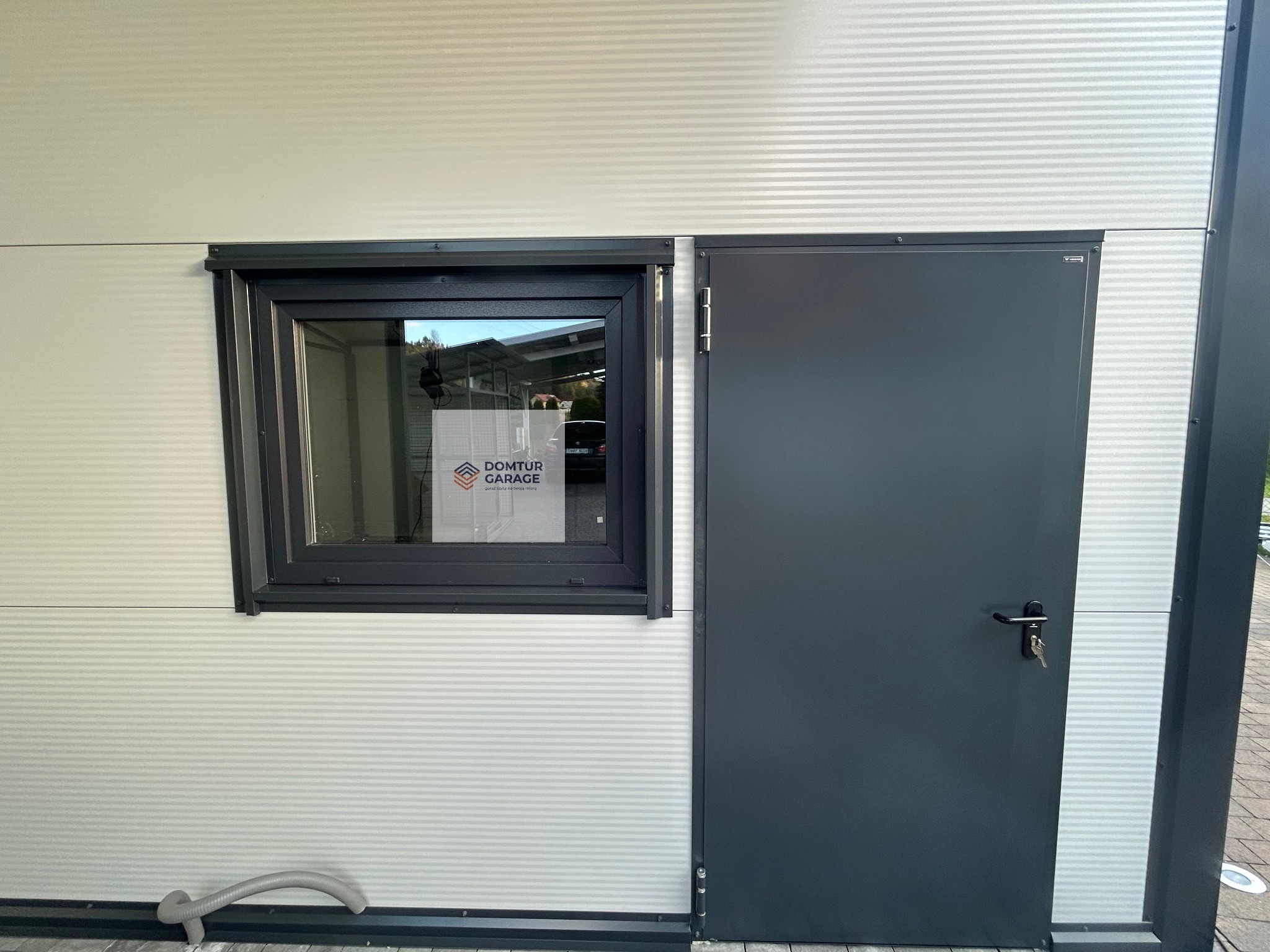 Nowoczesny garaż blaszany z oknem i drzwiami w Zagórzanach. Ściany z płyty warstwowej z widocznym ukrytym łącznikiem. W oknie widoczna kartka z logo firmy.
