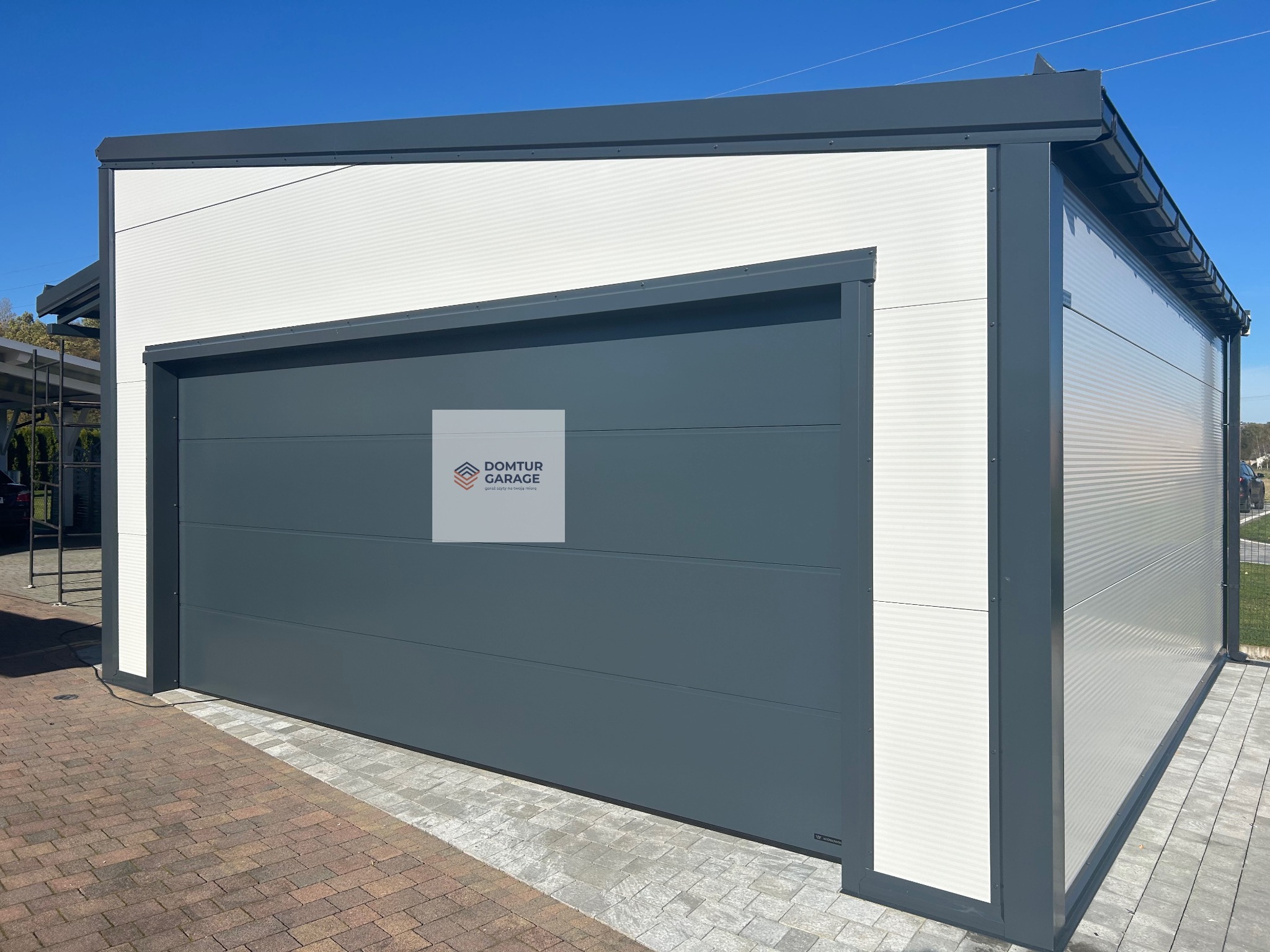 Nowoczesny garaż blaszany z szarą bramą segmentową i białymi ścianami z blachy trapezowej, widok z zewnątrz na tle kostki brukowej i błękitnego nieba.