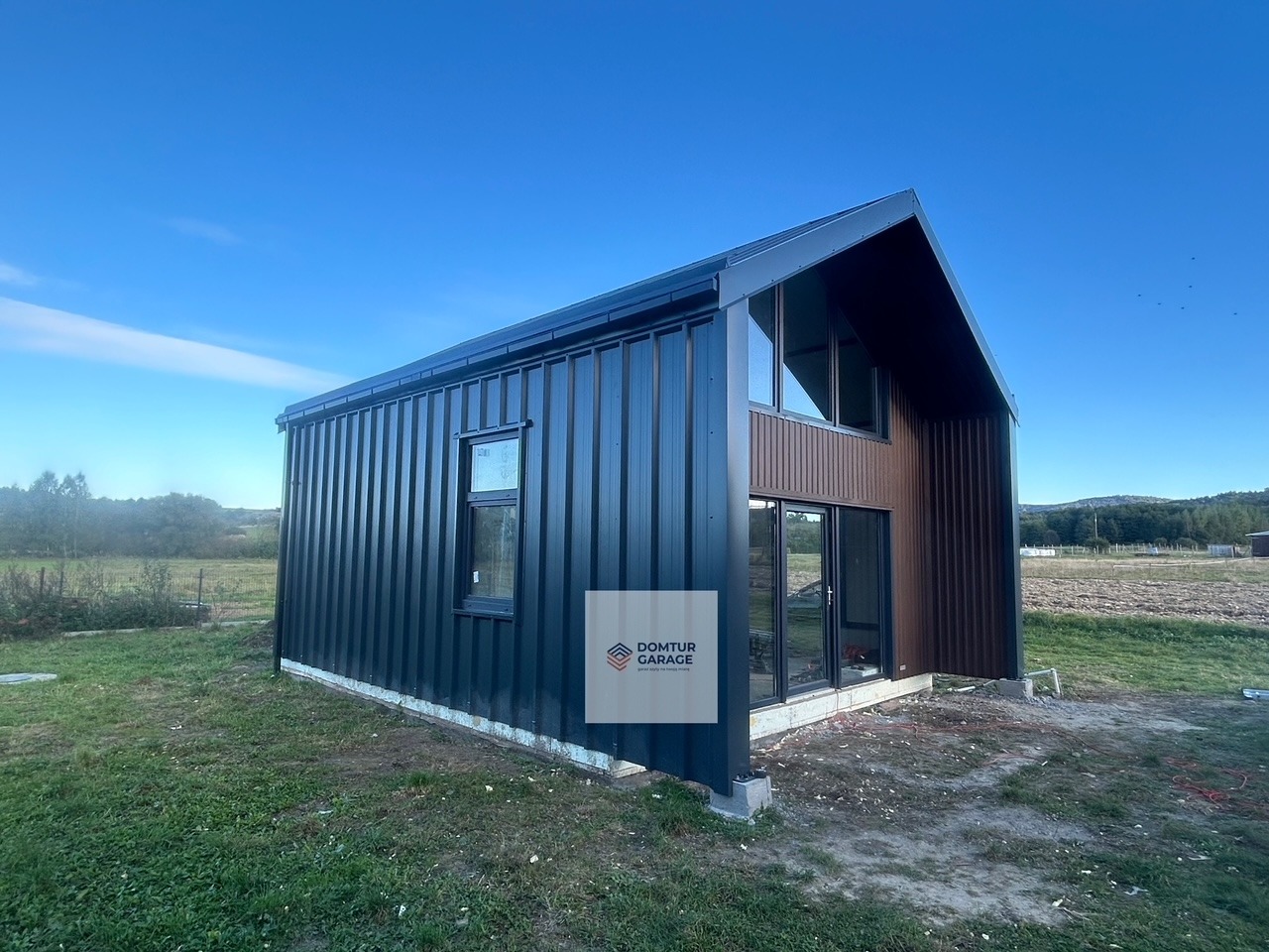 Nowoczesny, dwukolorowy garaż blaszany z oknami i drzwiami przesuwnymi, na fundamencie, w otoczeniu trawy i pola, pod błękitnym niebem.