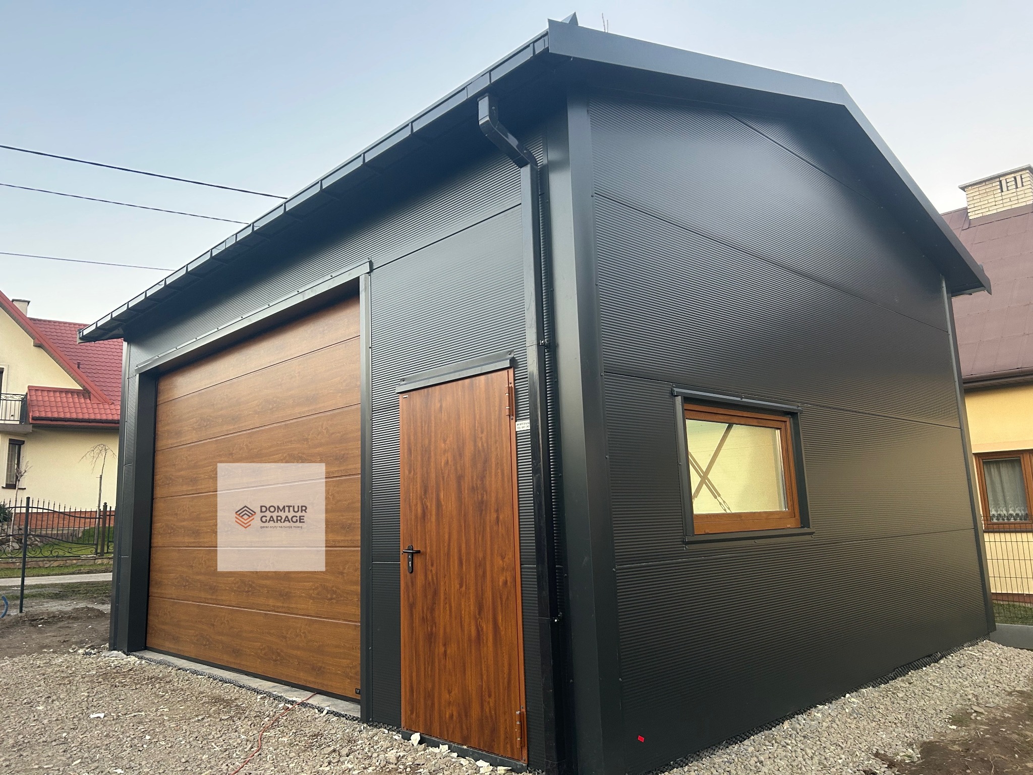 Czarny garaż blaszany z bramą segmentową w kolorze drewna i drzwiami wejściowymi. Ściany z płyty warstwowej z ukrytym łącznikiem. Widoczne okno i rynna.