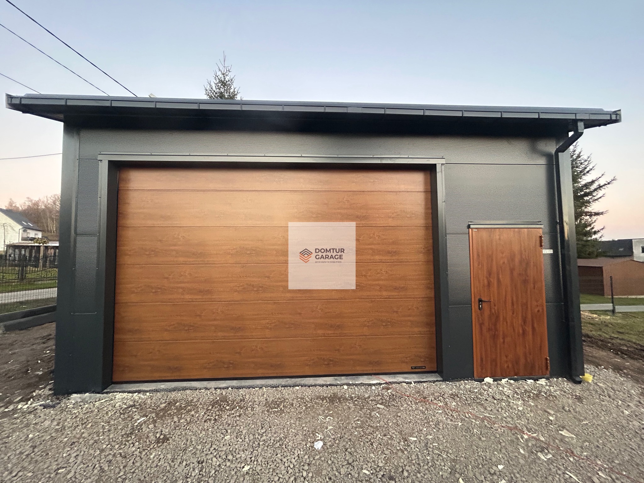 Nowoczesny garaż blaszany z brązową bramą segmentową i drzwiami wejściowymi, wykończony w kolorze grafitowym, na żwirowym podłożu.