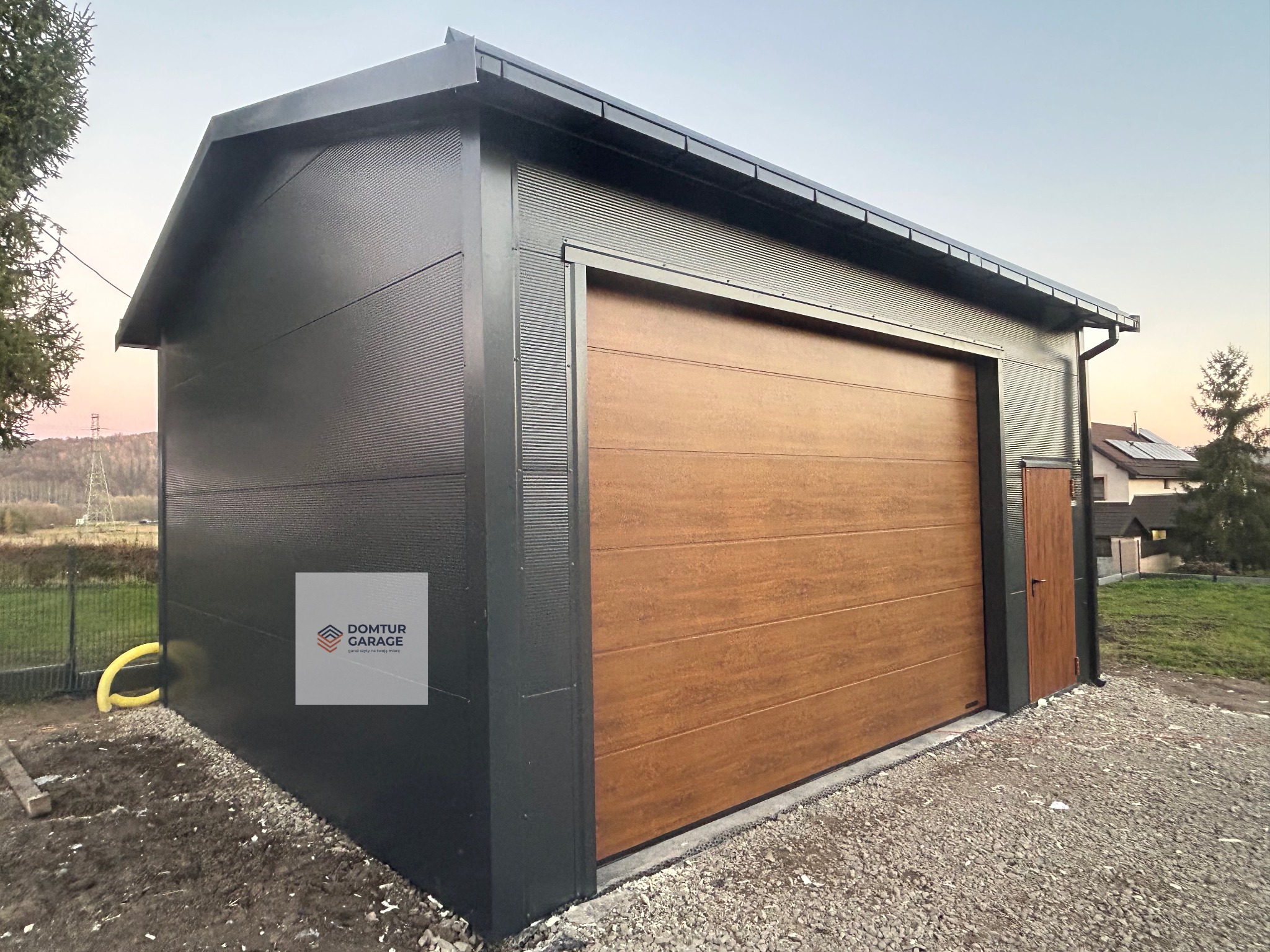 Nowoczesny garaż blaszany z brązową bramą segmentową i drzwiami wejściowymi, na żwirowym podłożu. Ściany garażu w kolorze grafitowym, ryflowane.