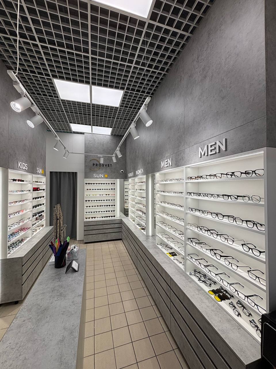 Wnętrze salonu optycznego z szarymi ścianami, oświetleniem sufitowym i ekspozycją okularów dla dzieci, kobiet i mężczyzn. Minimalistyczny design z akcentami kolorystycznymi.