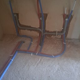 Hydrauliczn - Fragment instalacji wodnej z rur PEX w kolorach niebieskim i czerwonym, zamocowanych do ściany i podłogi. Widoczne szare rury kanalizacyjne w ścianie.
