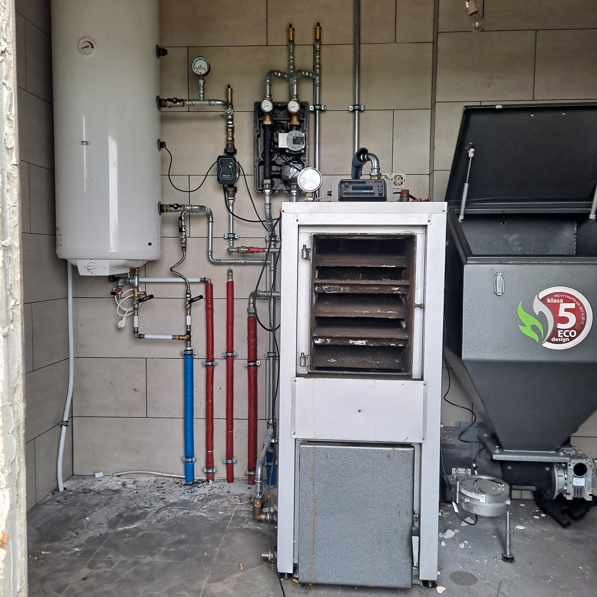 Kotłownia z piecem na ekogroszek klasy 5, zasobnikiem, bojlerem i instalacją CO z rurami w kolorze czerwonym i niebieskim. Widoczne manometry i sterownik.