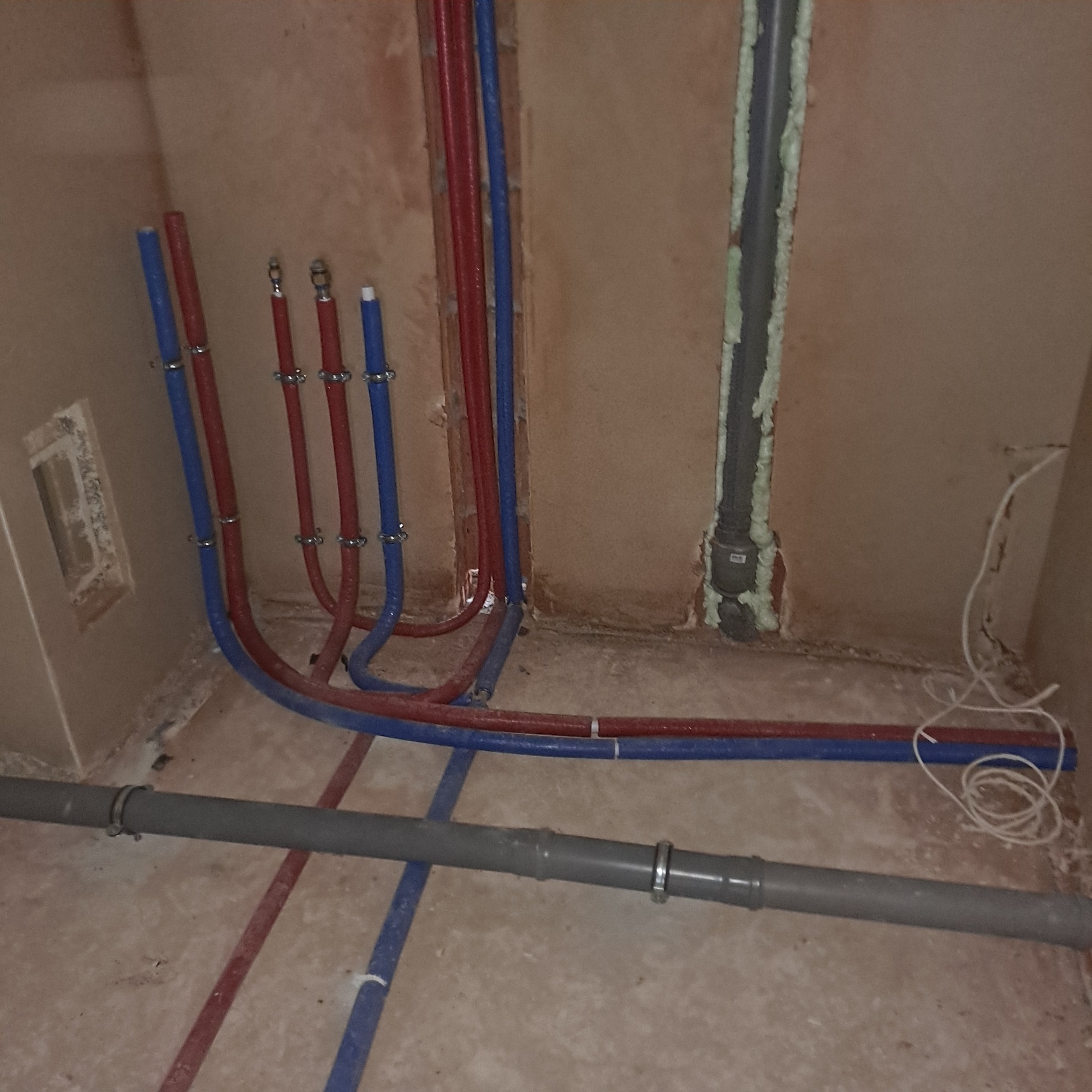 Instalacja wodna z rurami PEX (czerwone i niebieskie) zamocowanymi do ściany, szara rura kanalizacyjna na podłodze. Widoczne pianowanie wokół rury w ścianie.