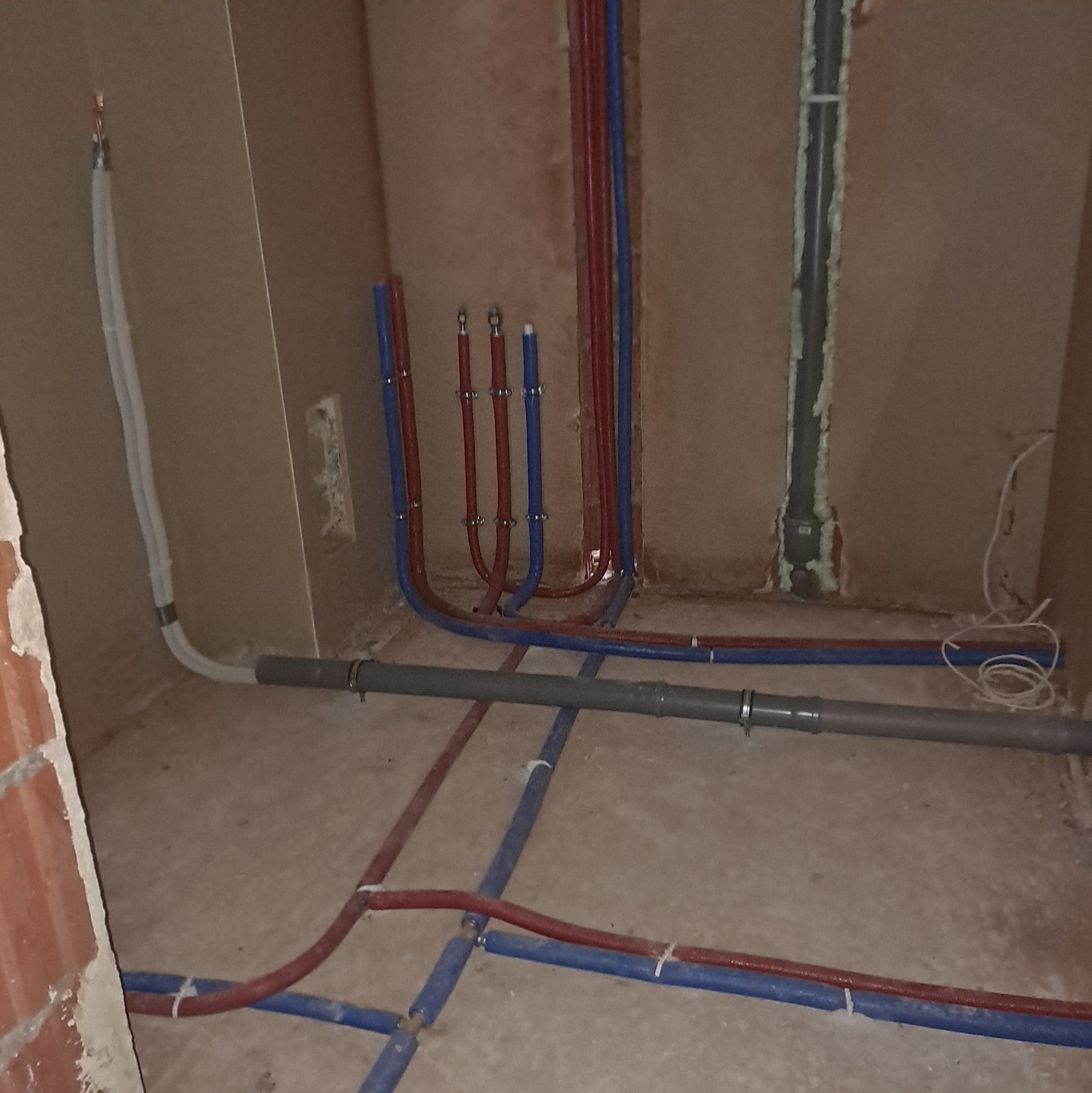 Wnętrze w trakcie instalacji wodno-kanalizacyjnej: widoczne rury w kolorach czerwonym i niebieskim, prowadzone po podłodze i w ścianach, z zamocowanymi uchwytami.