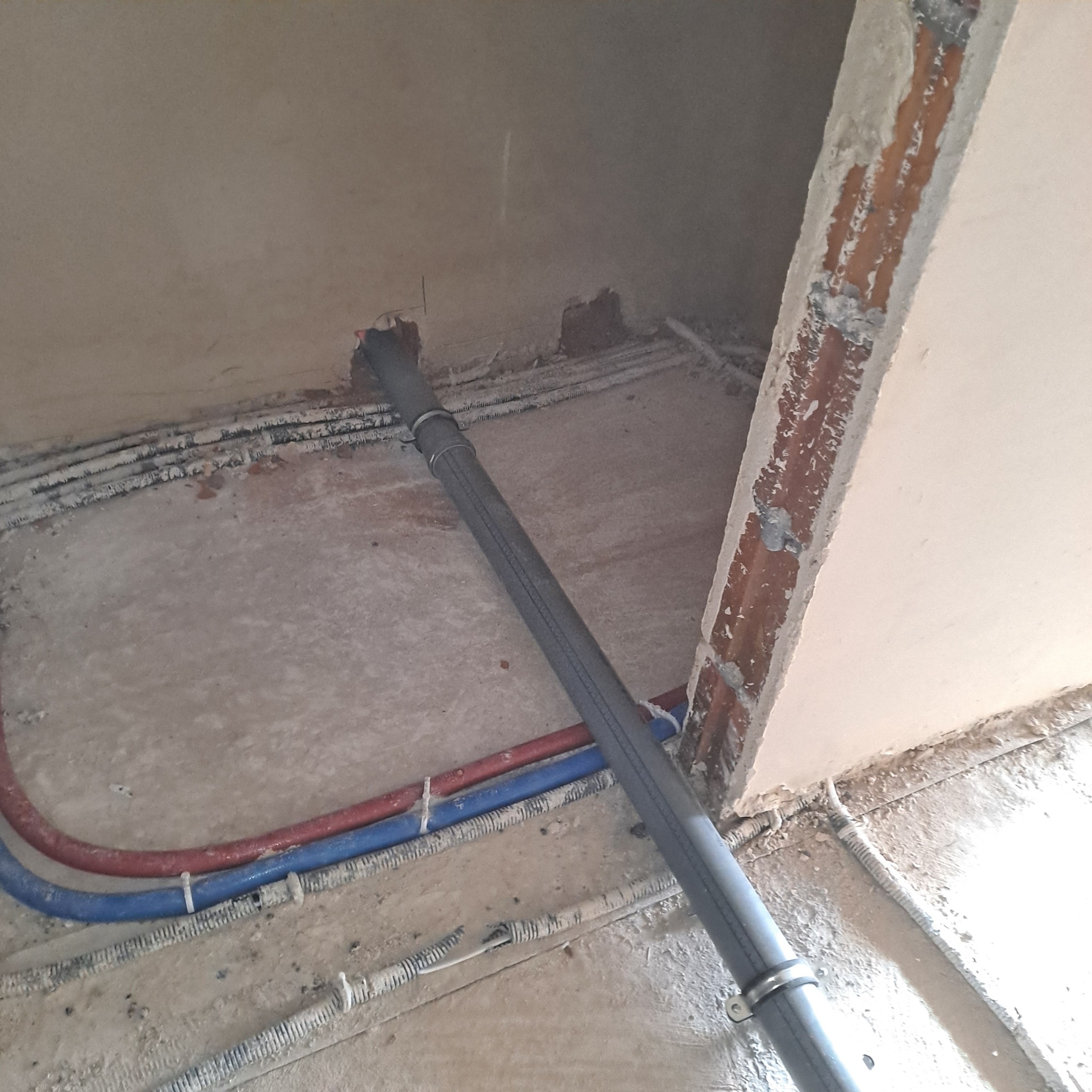 Szara rura kanalizacyjna z obejmami, ułożona na podłodze w surowym stanie, obok instalacji wodnej (czerwona i niebieska rura) i elektrycznej. Widoczne ślady po kuciu w ścianie.
