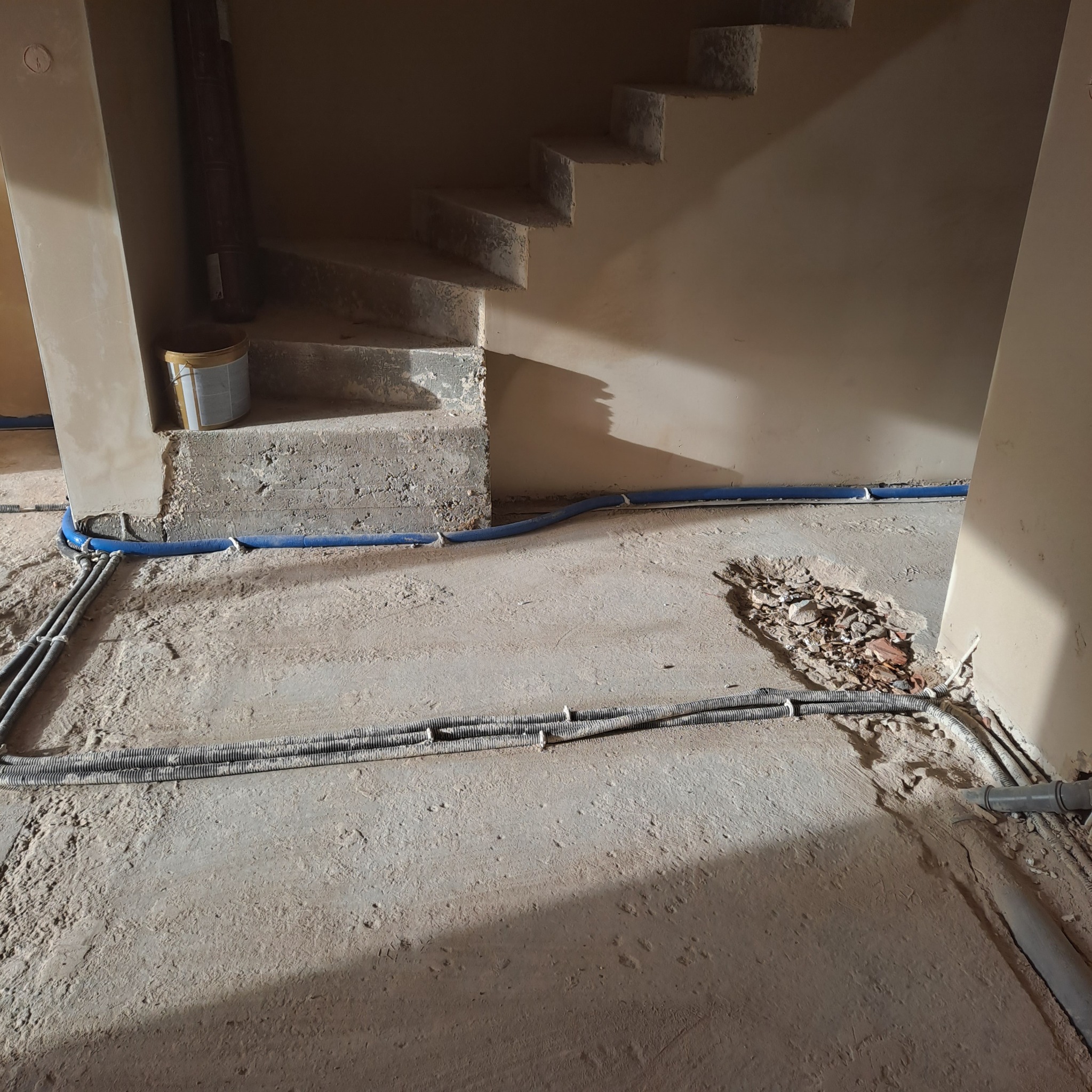 Instalacja rur i przewodów w surowym wnętrzu z betonową podłogą i schodami. Widoczne rury w kolorze niebieskim i szare peszle. Etap prac wykończeniowych.