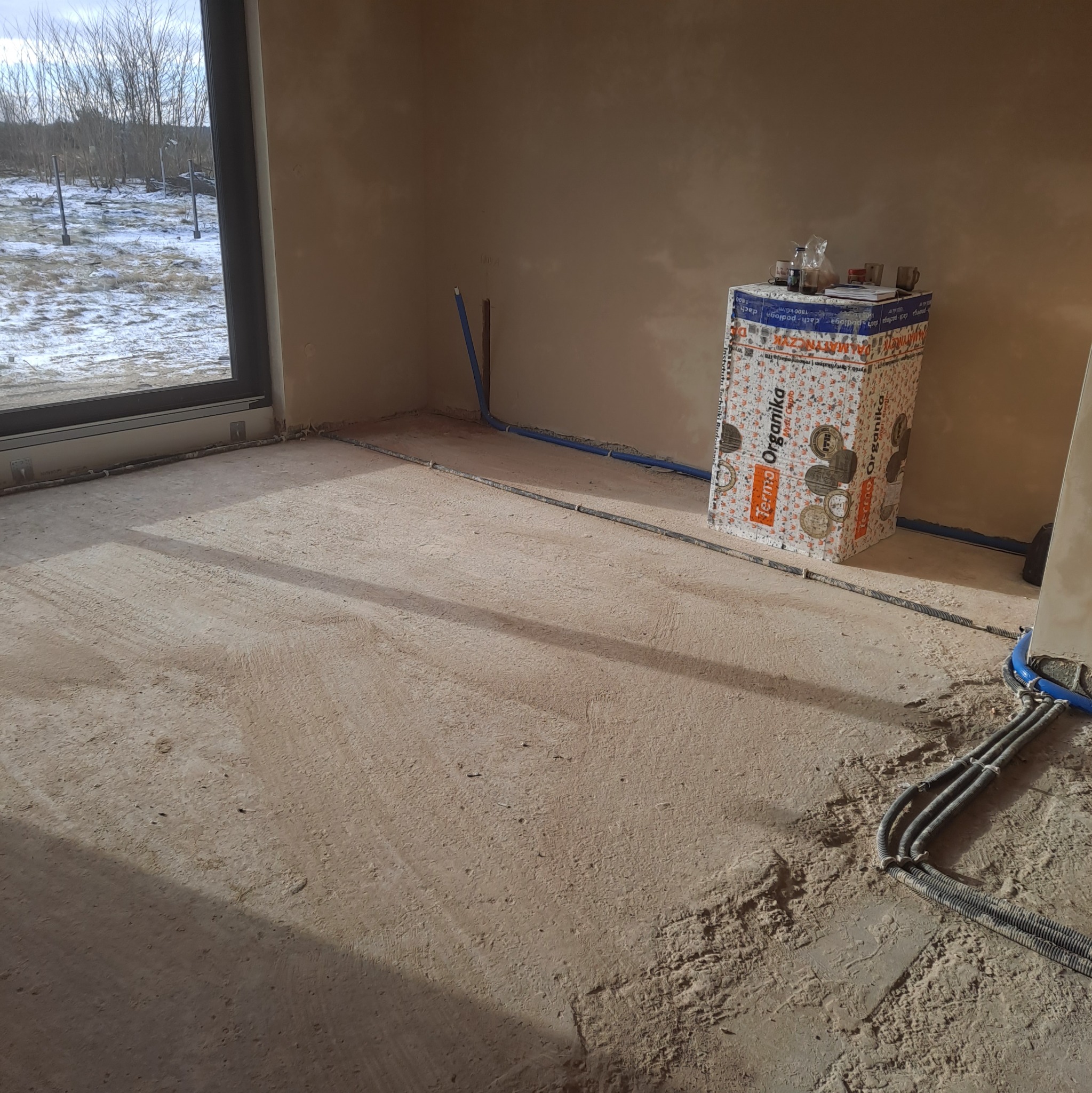 Wnętrze w trakcie prac wykończeniowych: widoczne rury instalacji podłogowej, styropian Termo Organika i surowe ściany. Okno z widokiem na zimowy krajobraz.