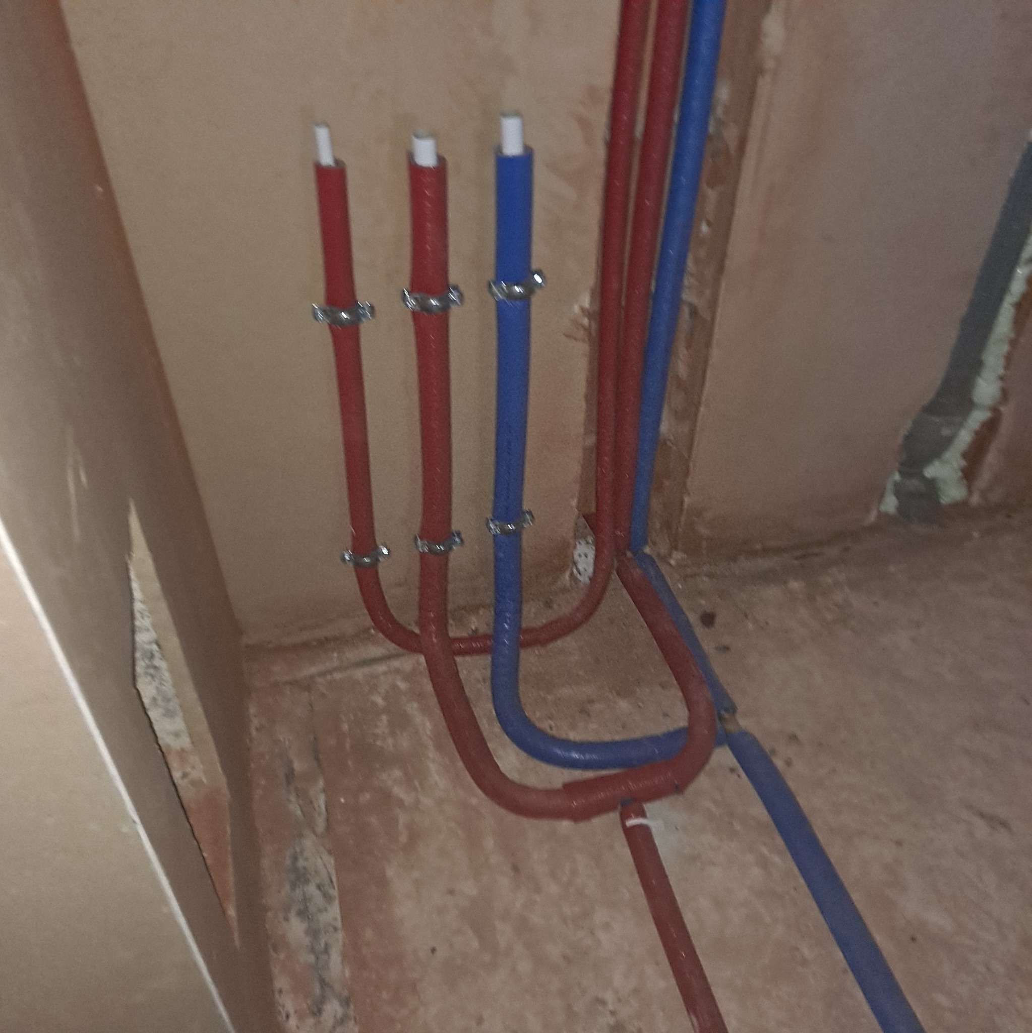 Czerwone i niebieskie rury instalacji wodnej, przymocowane metalowymi uchwytami do ściany w surowym stanie. Widoczne fragmenty podłogi i ściany.