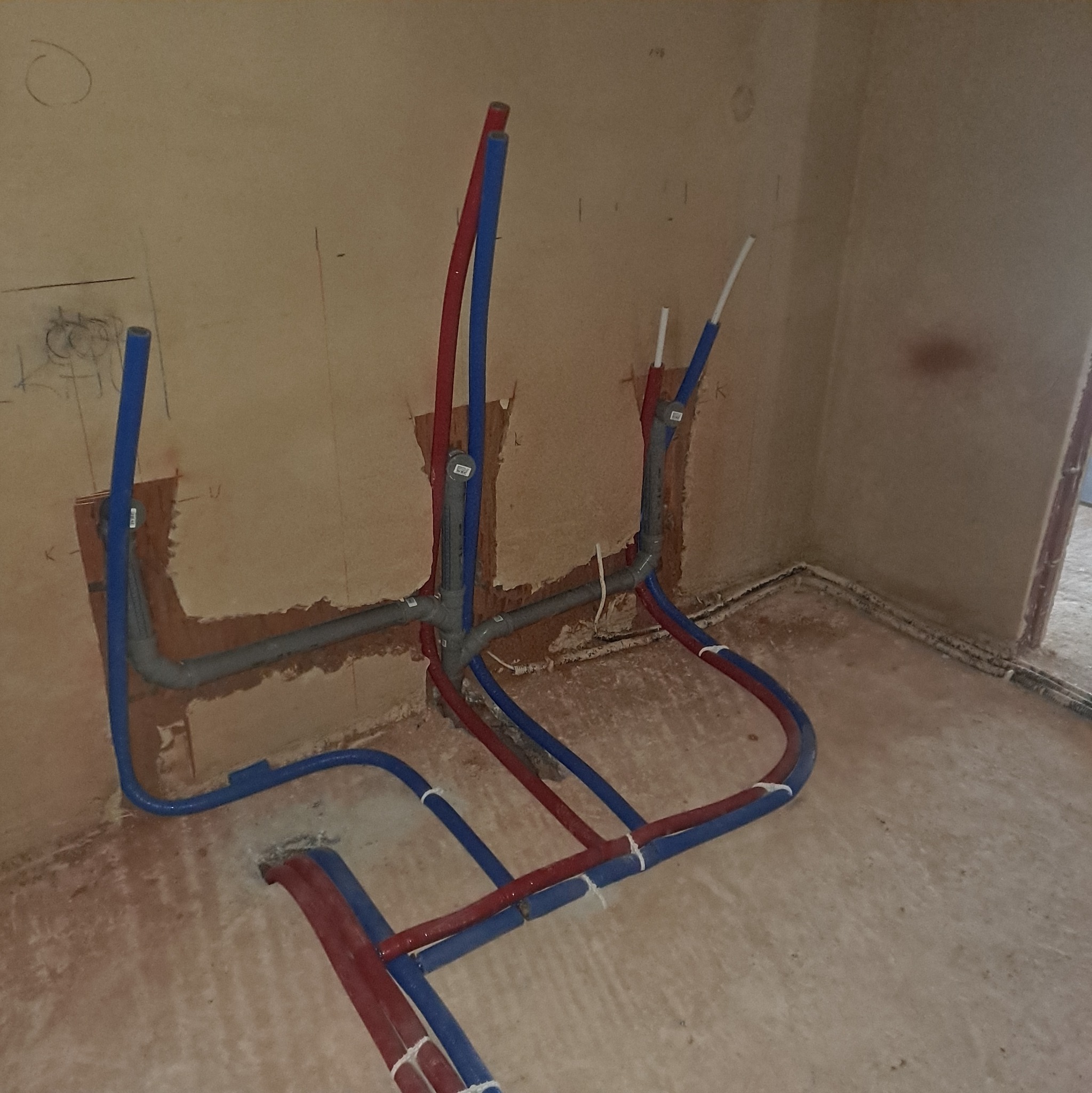 Instalacja wodna i grzewcza w surowym wnętrzu. Rury PEX w kolorach czerwonym i niebieskim, częściowo zamontowane w ścianie. Widoczne łączenia i mocowania.