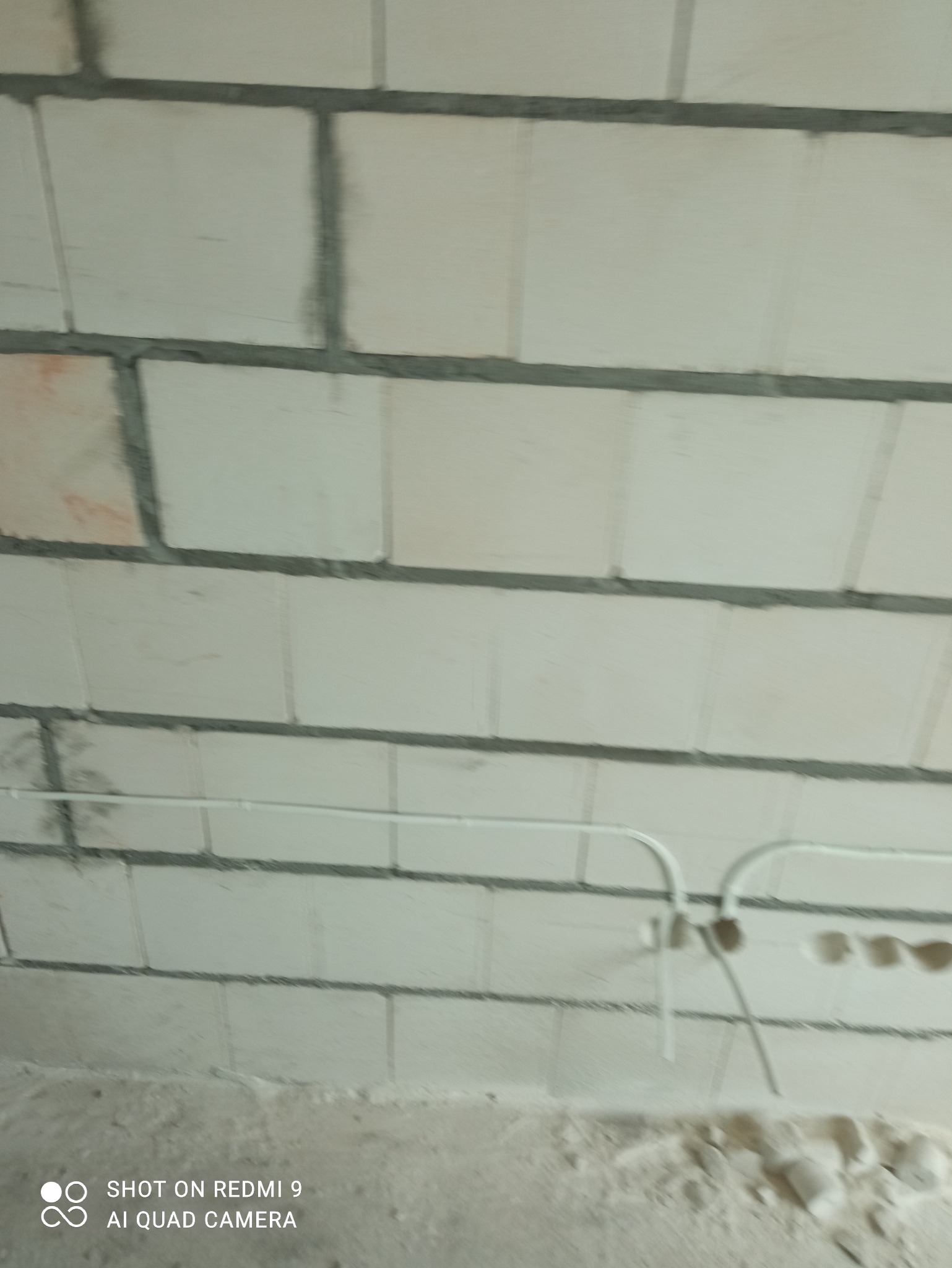 Biała ściana z betonu komórkowego z widoczną, białą instalacją elektryczną w rurkach. Widoczne otwory pod gniazdka. Surowy stan wykończenia.