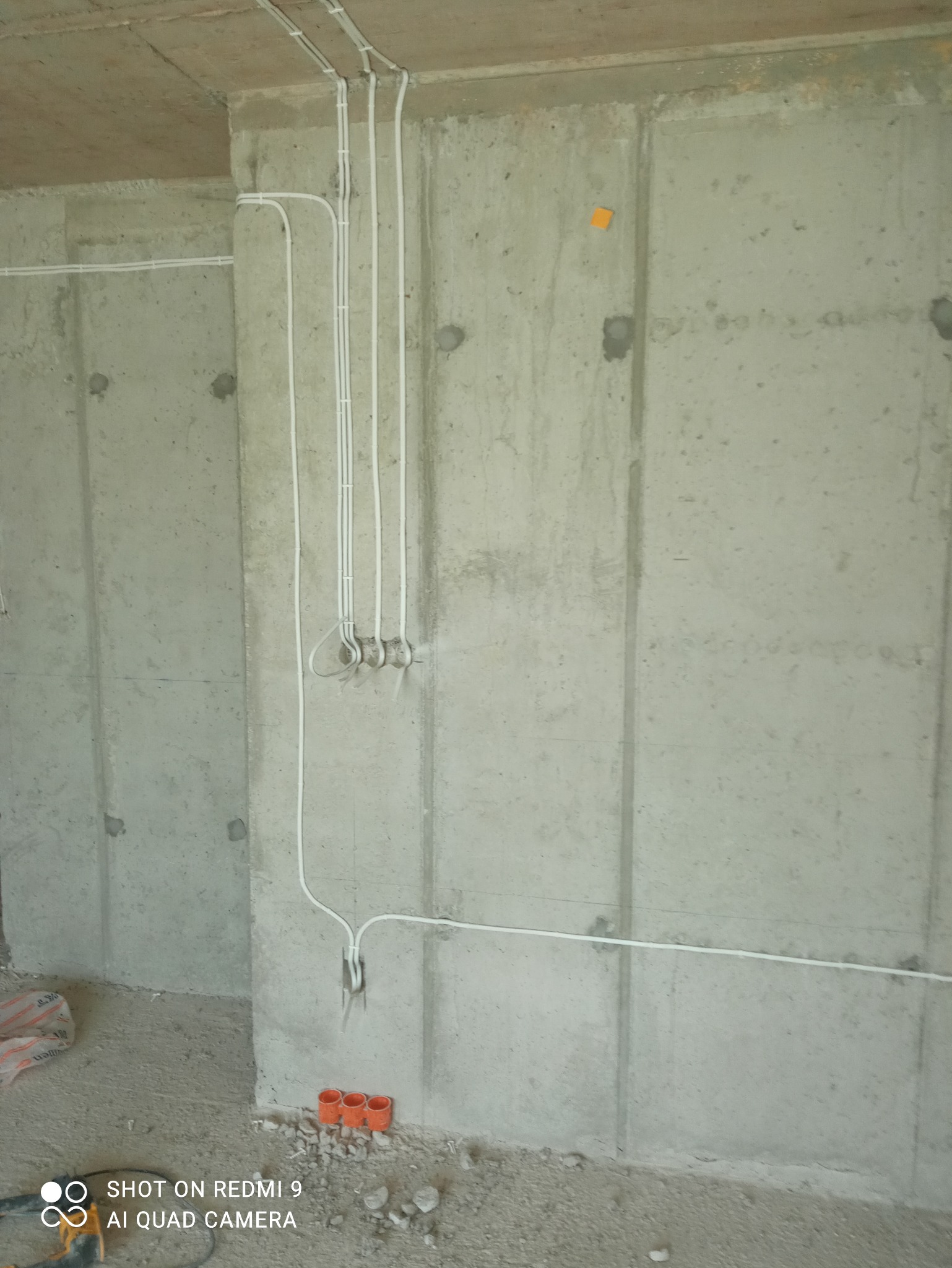 Surowa ściana z betonu z widoczną, częściowo zamontowaną instalacją elektryczną w białych rurkach. Na podłodze leżą pomarańczowe puszki instalacyjne.