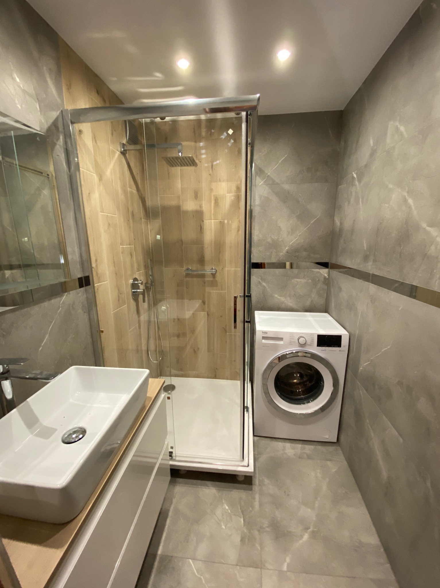 Nowoczesna łazienka z kabiną prysznicową typu walk-in, umywalką nablatową i pralką. Szare płytki imitujące marmur, drewnopodobne wykończenie w kabinie.