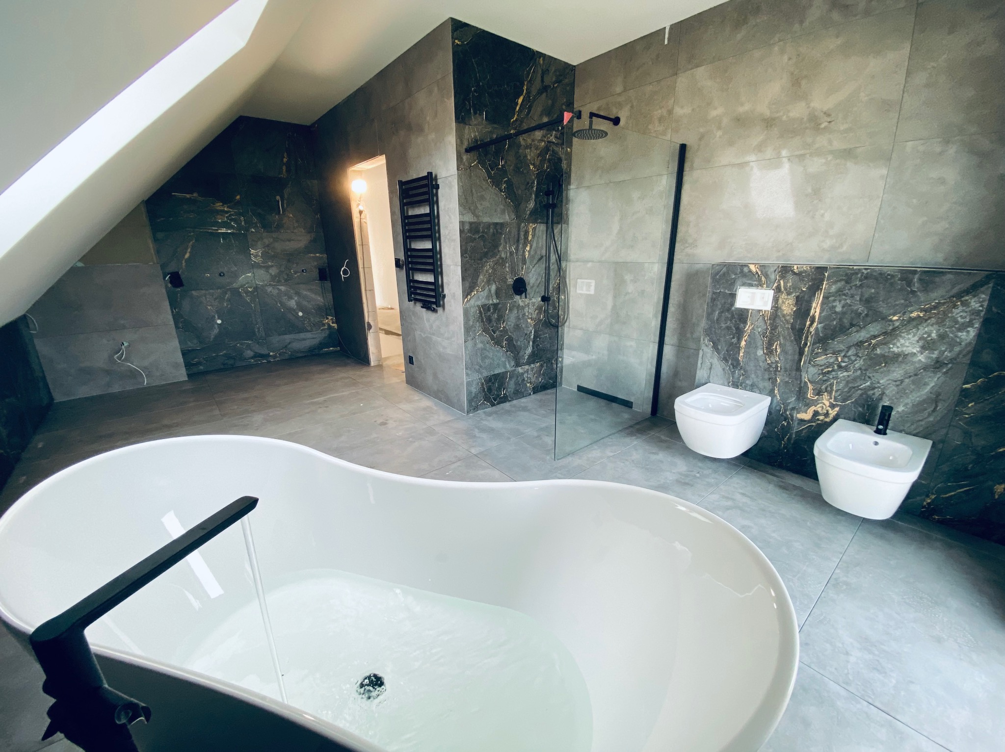 Nowoczesna łazienka na poddaszu z wolnostojącą wanną wypełnioną wodą, kabiną prysznicową typu walk-in i marmurowymi płytkami w odcieniach szarości i czerni.