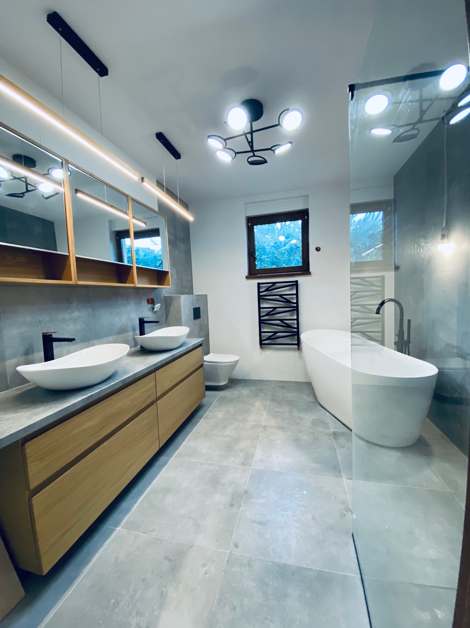 Nowoczesna łazienka z dwoma umywalkami na drewnianej szafce, wolnostojąca wanna za szklaną ścianką i szara płytka podłogowa. Minimalistyczny design.