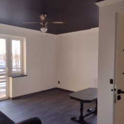 MMJ Marcin Jakubas - Pokój po malowaniu: ciemny sufit z wentylatorem, białe ściany, okno balkonowe i ciemna podłoga. Widoczny fragment sofy i stolika. Nowoczesne wykończenie wnętrza.