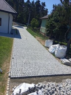 Ułożona kostka brukowa na podjeździe do domu, widok z góry. Częściowo ułożona kostka, worki z materiałem budowlanym w tle. Widoczne drzewa i budynek.