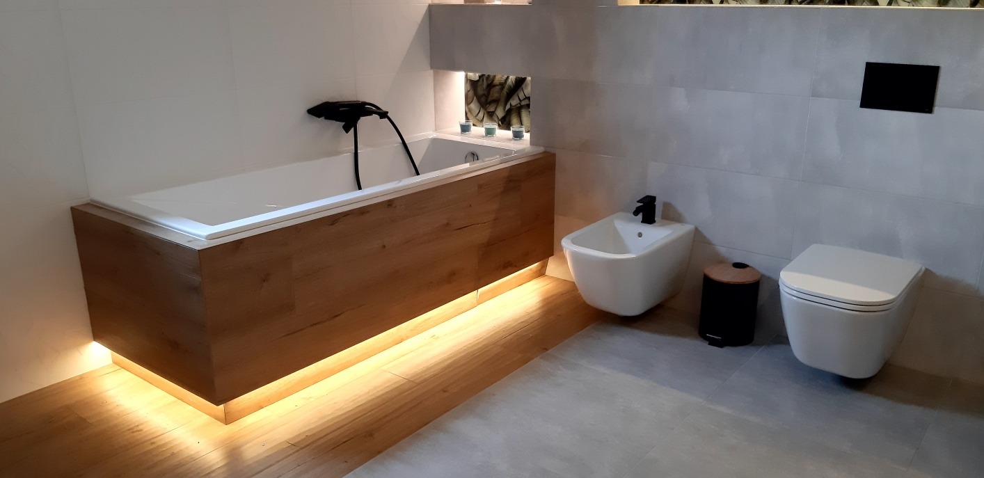 Nowoczesna łazienka z podświetlaną wanną obudowaną drewnem, bidetem i podwieszaną toaletą. Szare płytki na ścianach i podłodze. Minimalistyczny design.