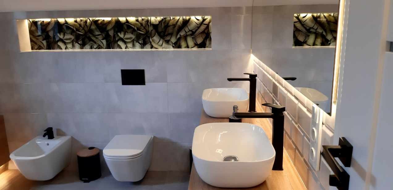 Nowoczesna łazienka z dwoma białymi umywalkami, czarną armaturą i podświetlanym panelem dekoracyjnym nad toaletą i bidetem. Minimalistyczny design.