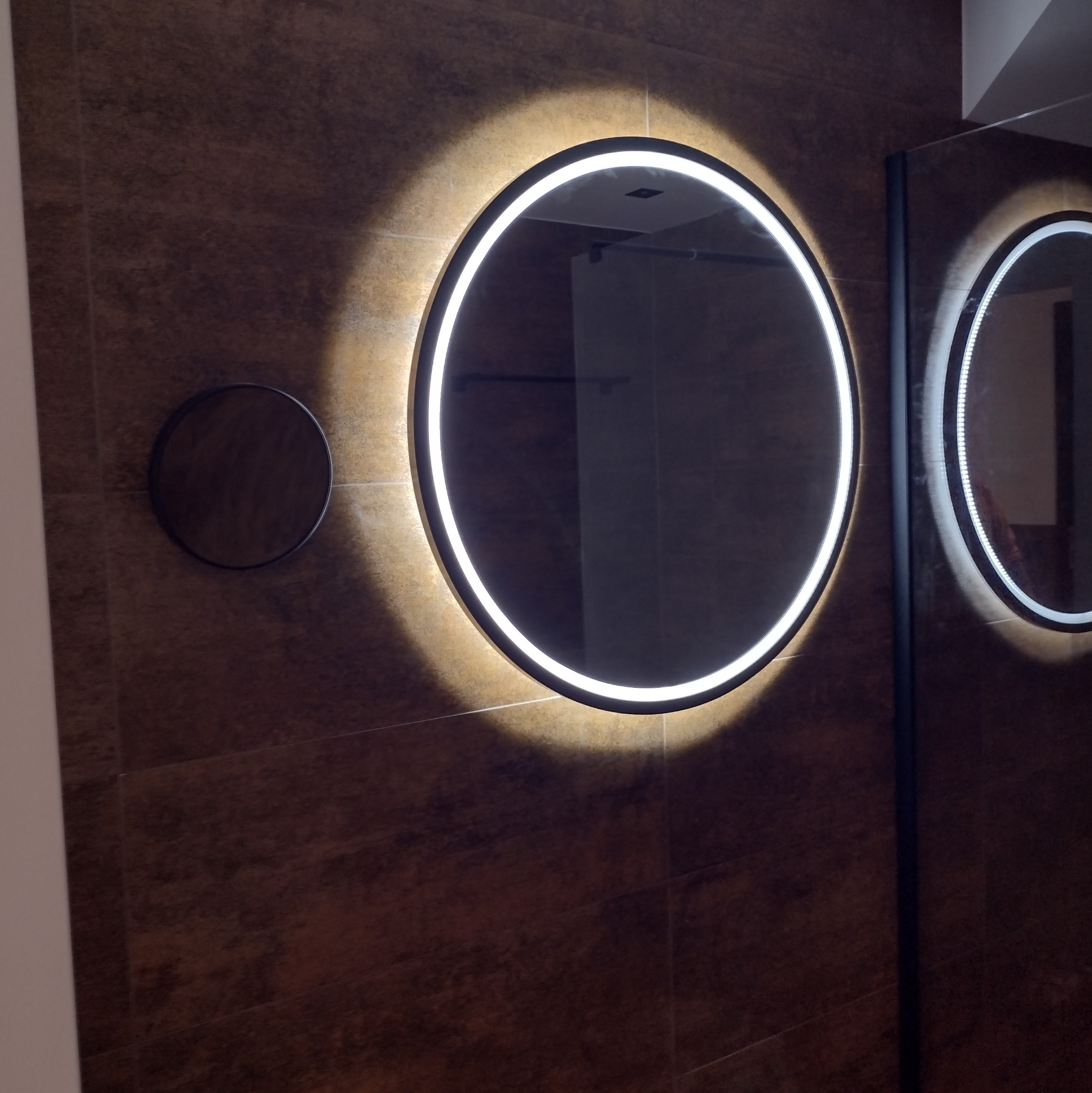 Nowoczesna łazienka z dwoma okrągłymi lustrami LED na brązowej ścianie z płytek. Dodatkowe małe okrągłe lustro po lewej stronie. Minimalistyczny design.