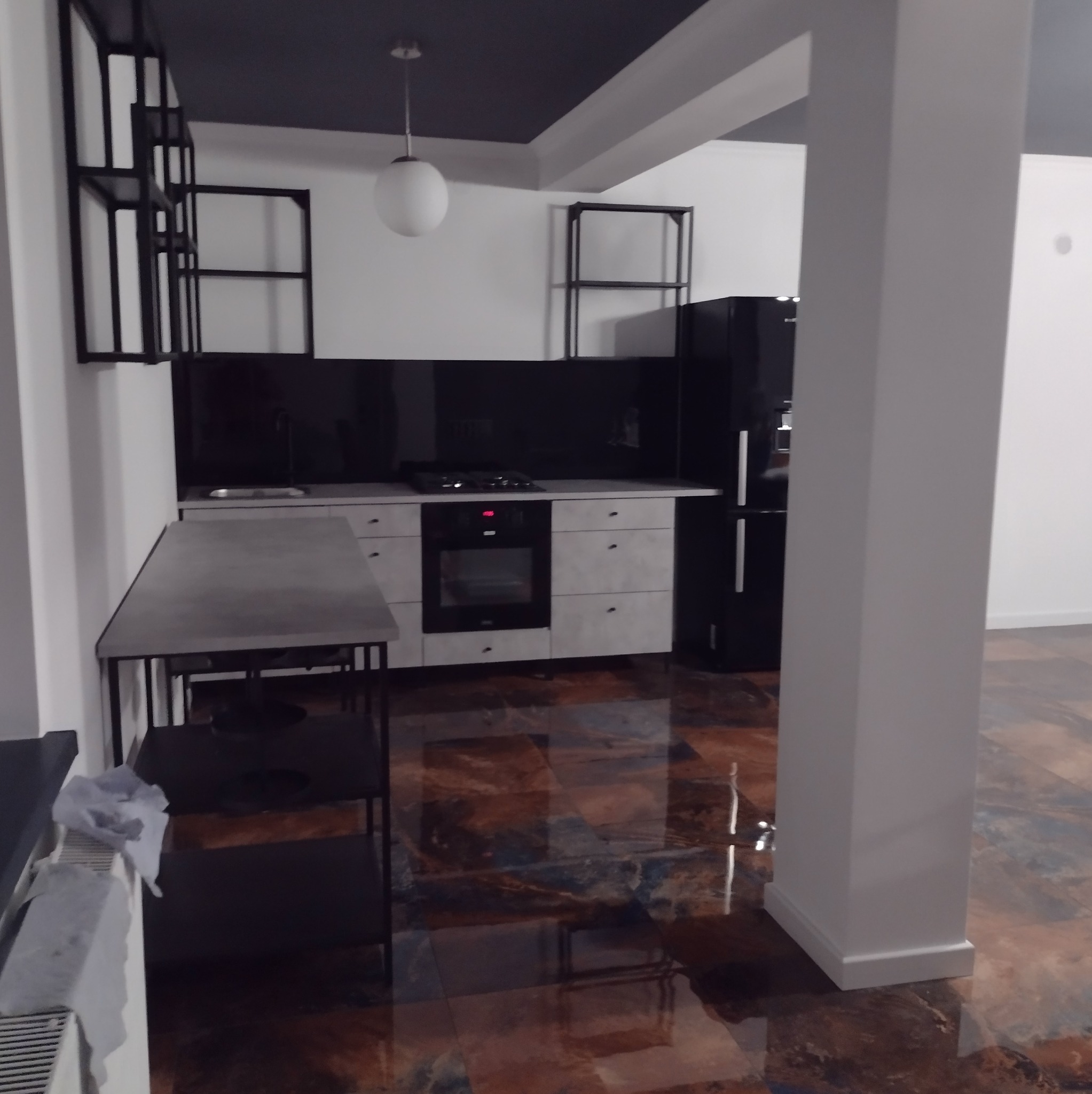 Nowoczesna kuchnia z czarnymi akcentami: szafki w odcieniach szarości, czarny blat i lodówka. Podłoga z efektem żywicy w odcieniach brązu. Minimalistyczne półki.