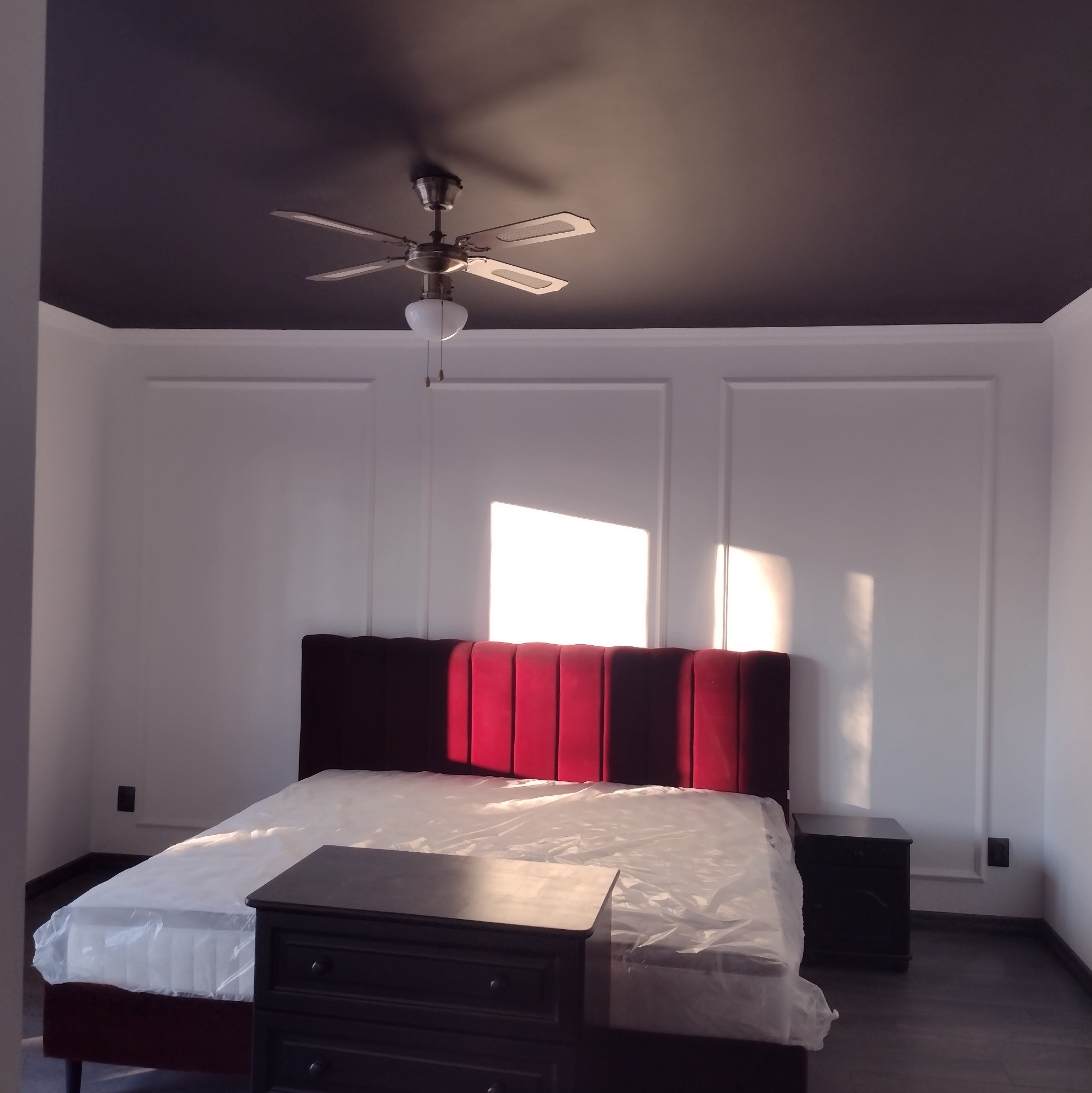 Nowoczesna sypialnia z ciemnym sufitem i dekoracyjnymi panelami ściennymi. Czerwone wezgłowie łóżka i szafki nocne w ciemnym odcieniu. Widoczny wentylator sufitowy.