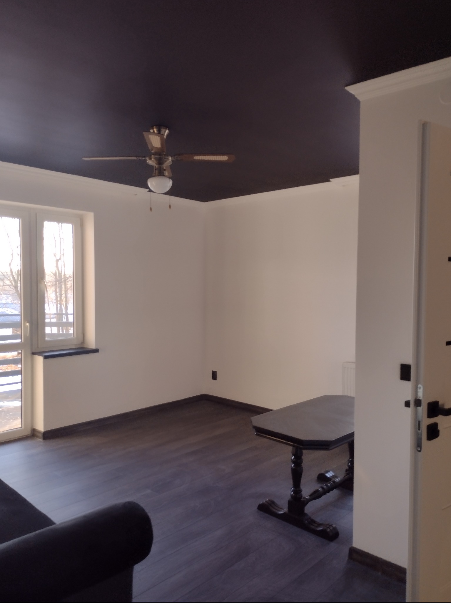 Pokój po malowaniu: ciemny sufit z wentylatorem, białe ściany, okno balkonowe i ciemna podłoga. Widoczny fragment sofy i stolika. Nowoczesne wykończenie wnętrza.
