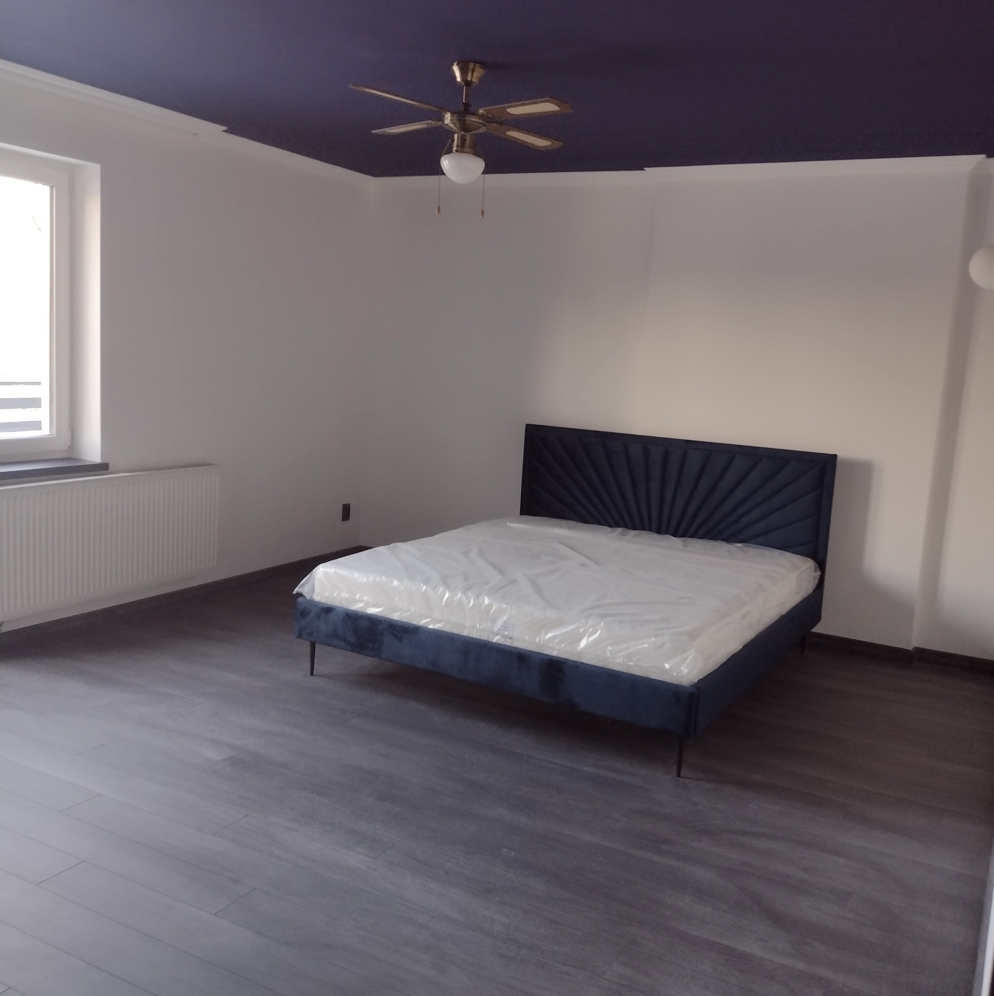 Pokój po remoncie: ciemna podłoga, białe ściany, granatowy sufit i łóżko z zagłówkiem. Widoczny wentylator sufitowy. Minimalistyczny design.