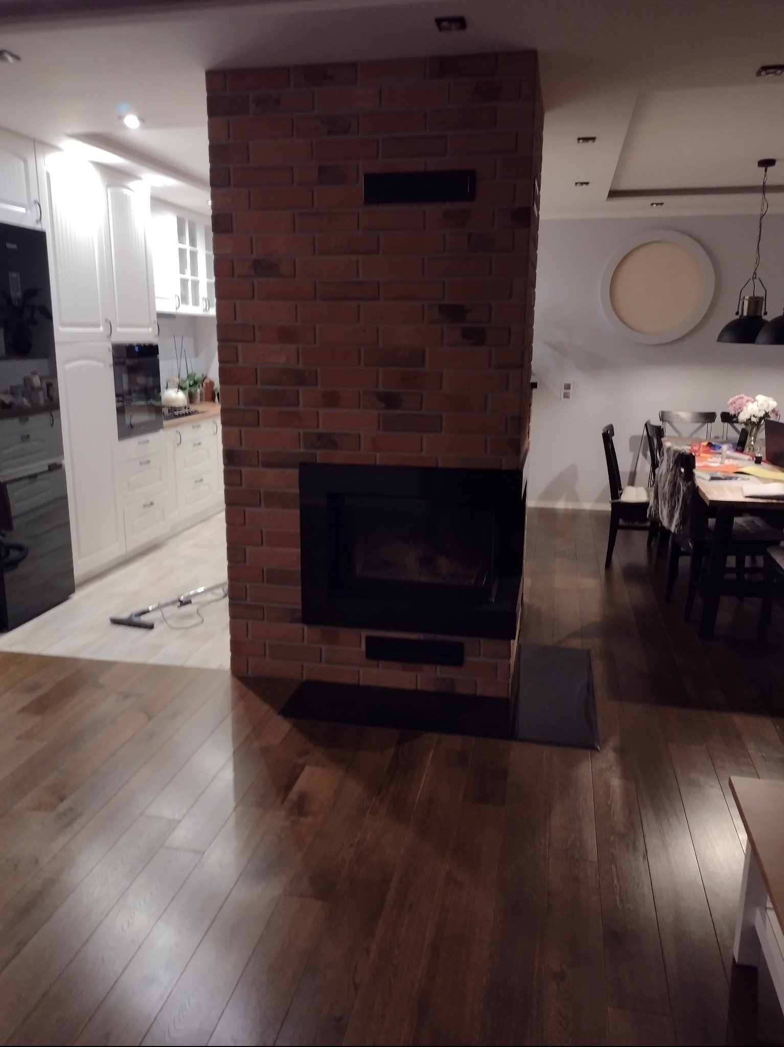 Ceglany kominek z czarnym wkładem w nowoczesnym wnętrzu. Parkiet na podłodze, w tle biała kuchnia i jadalnia z okrągłym elementem dekoracyjnym na ścianie.