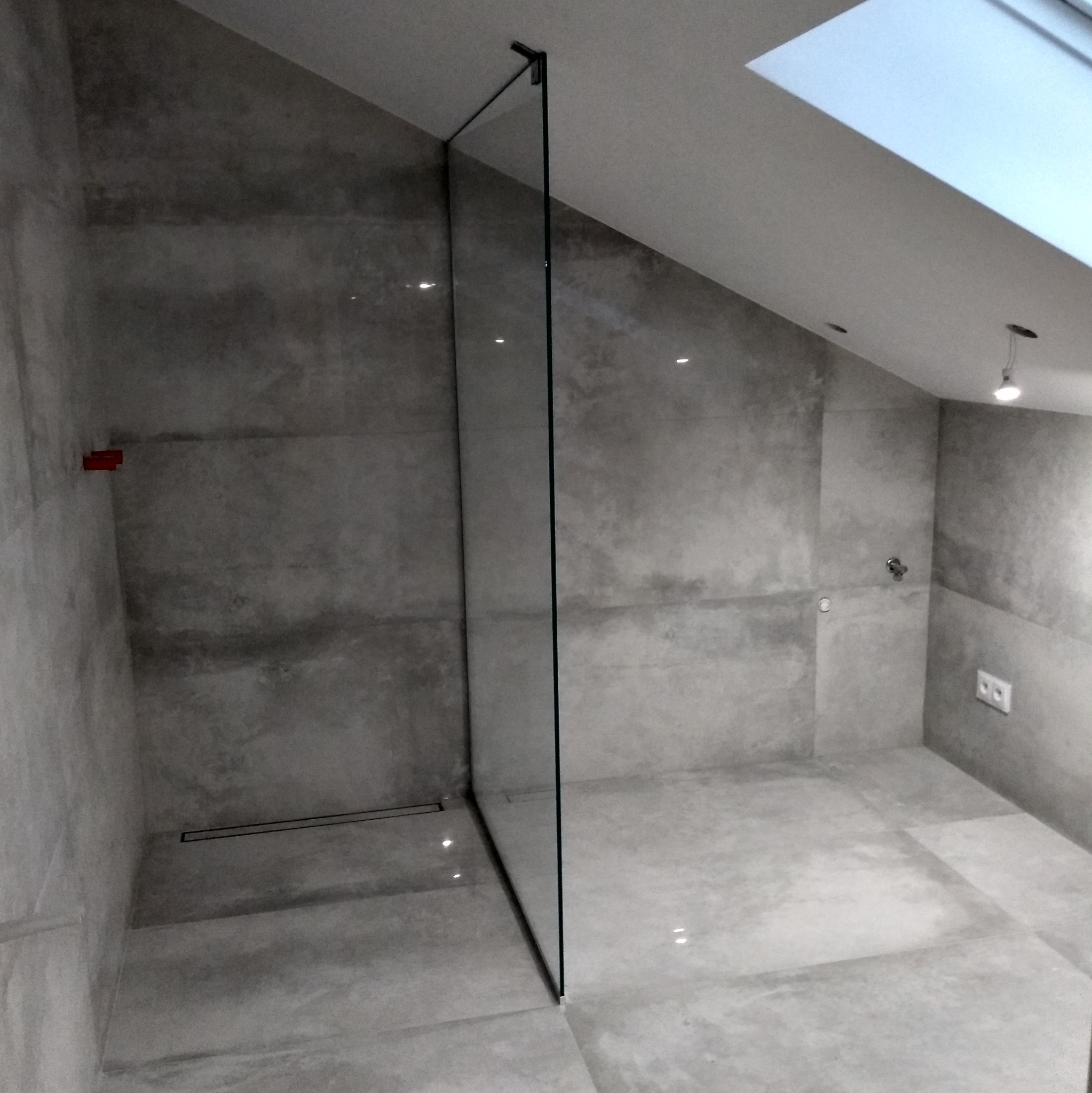 Nowoczesna łazienka w odcieniach szarości z kabiną prysznicową typu walk-in i odpływem liniowym. Uwagę zwraca skośny sufit z oknem dachowym.