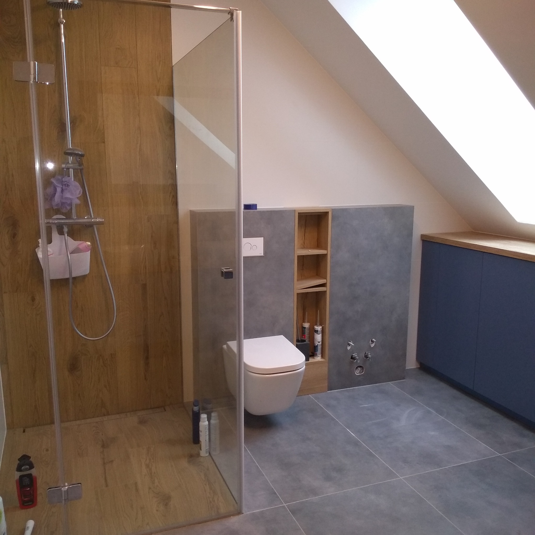 Nowoczesna łazienka na poddaszu z kabiną prysznicową typu walk-in, wiszącą toaletą i zabudową meblową w odcieniach szarości, drewna i granatu. Uwagę zwraca skośny sufit.