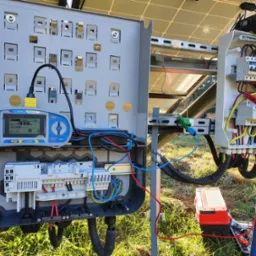 Szary rozdzielacz elektryczny z podłączonym miernikiem rezystancji izolacji Metrel EurotestPV, widoczne panele słoneczne w tle oraz skrzynka z bezpiecznikami i przewodami.