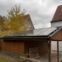 Drewniany garaż z panelami słonecznymi na dachu, widok z zewnątrz w pochmurny dzień, w tle domy i drzewa z jesiennymi liśćmi.