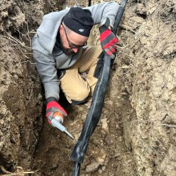 Elektrojar Usługi Elektryczne i Fotowoltaika - Elektryk w wykopie łączy kabel wysokiego napięcia za pomocą specjalistycznego pistoletu. Praca w trudnych warunkach gruntowych, widoczne warstwy ziemi.