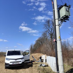 Elektrojar Usługi Elektryczne i Fotowoltaika - Biały samochód dostawczy Toyota obok betonowego słupa energetycznego z transformatorem, widoczne prace ziemne i instalacja kabli. Błękitne niebo z chmurami w tle.