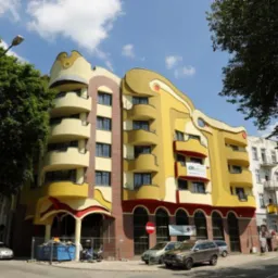 Fasada budynku o unikalnym, falistym designie w odcieniach żółci i czerwieni, z widocznymi balkonami, otoczona zielenią drzew i zaparkowanymi samochodami.