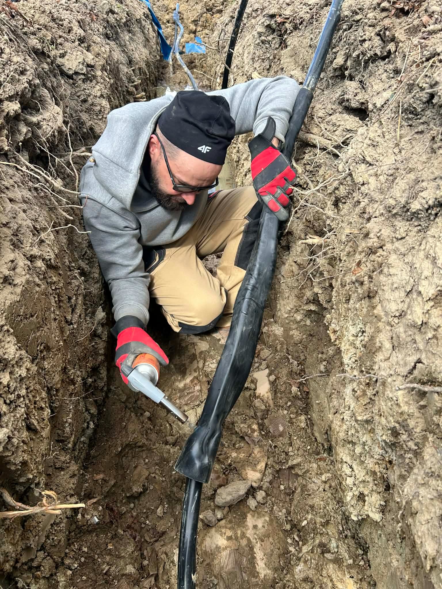 Elektryk w wykopie łączy kabel wysokiego napięcia za pomocą specjalistycznego pistoletu. Praca w trudnych warunkach gruntowych, widoczne warstwy ziemi.