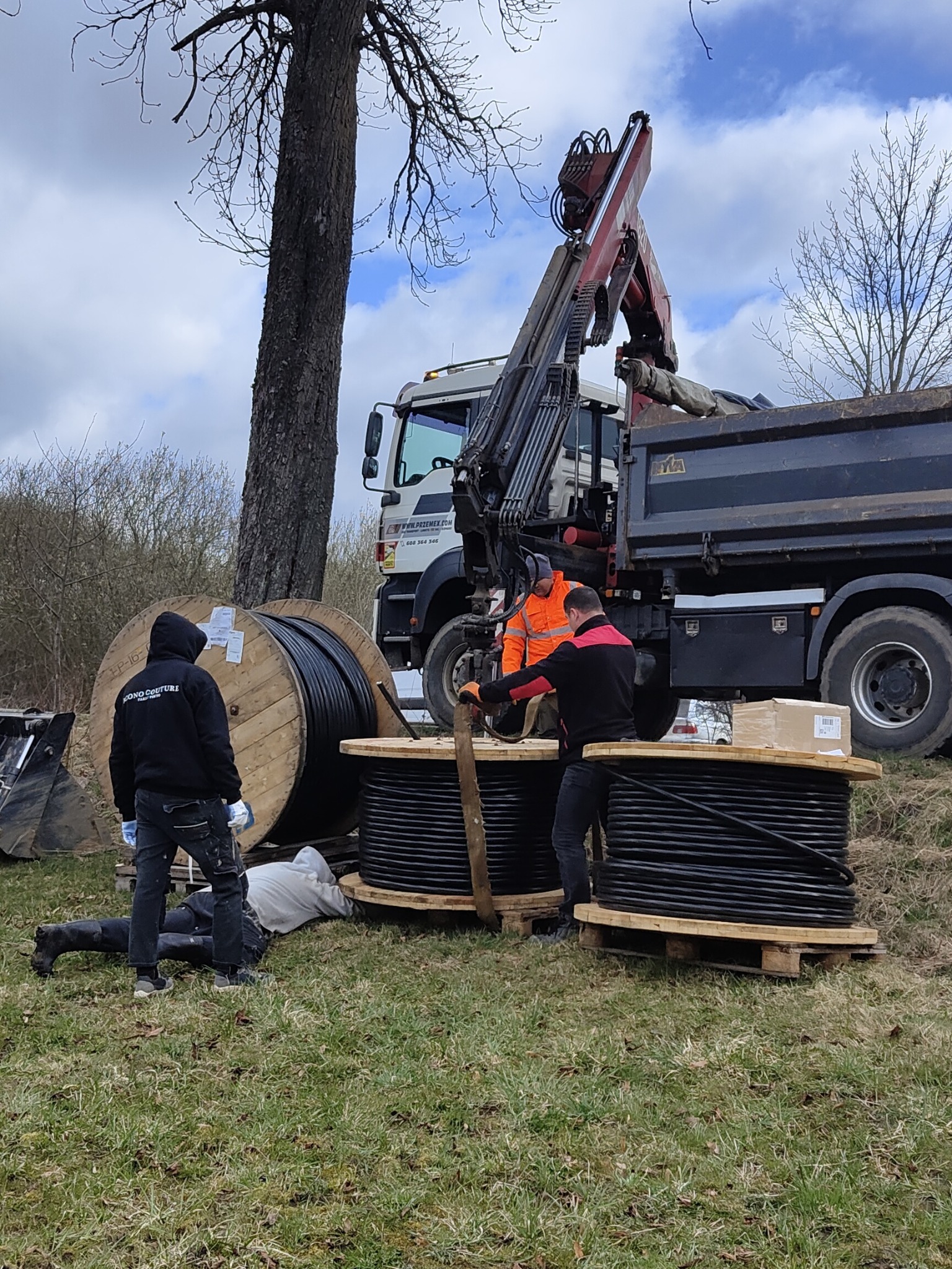Prace ziemne związane z układaniem kabli: robotnicy przy bębnach z grubym, czarnym kablem, ciężarówka z dźwigiem w tle, pochmurne niebo.