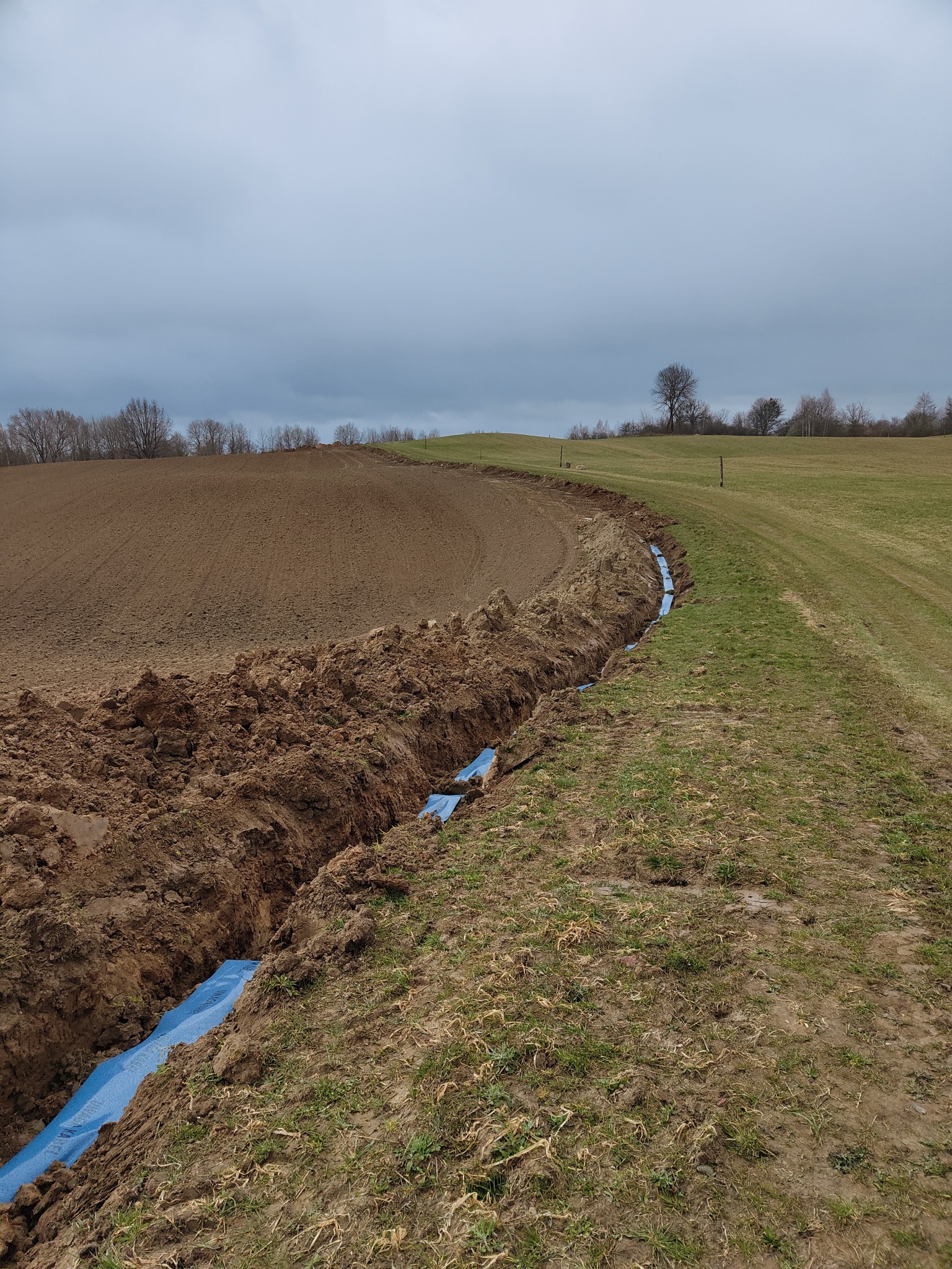 Wykopany rów w ziemi z niebieską rurą w środku, prawdopodobnie do instalacji podziemnej. Widok na pole i pochmurne niebo.