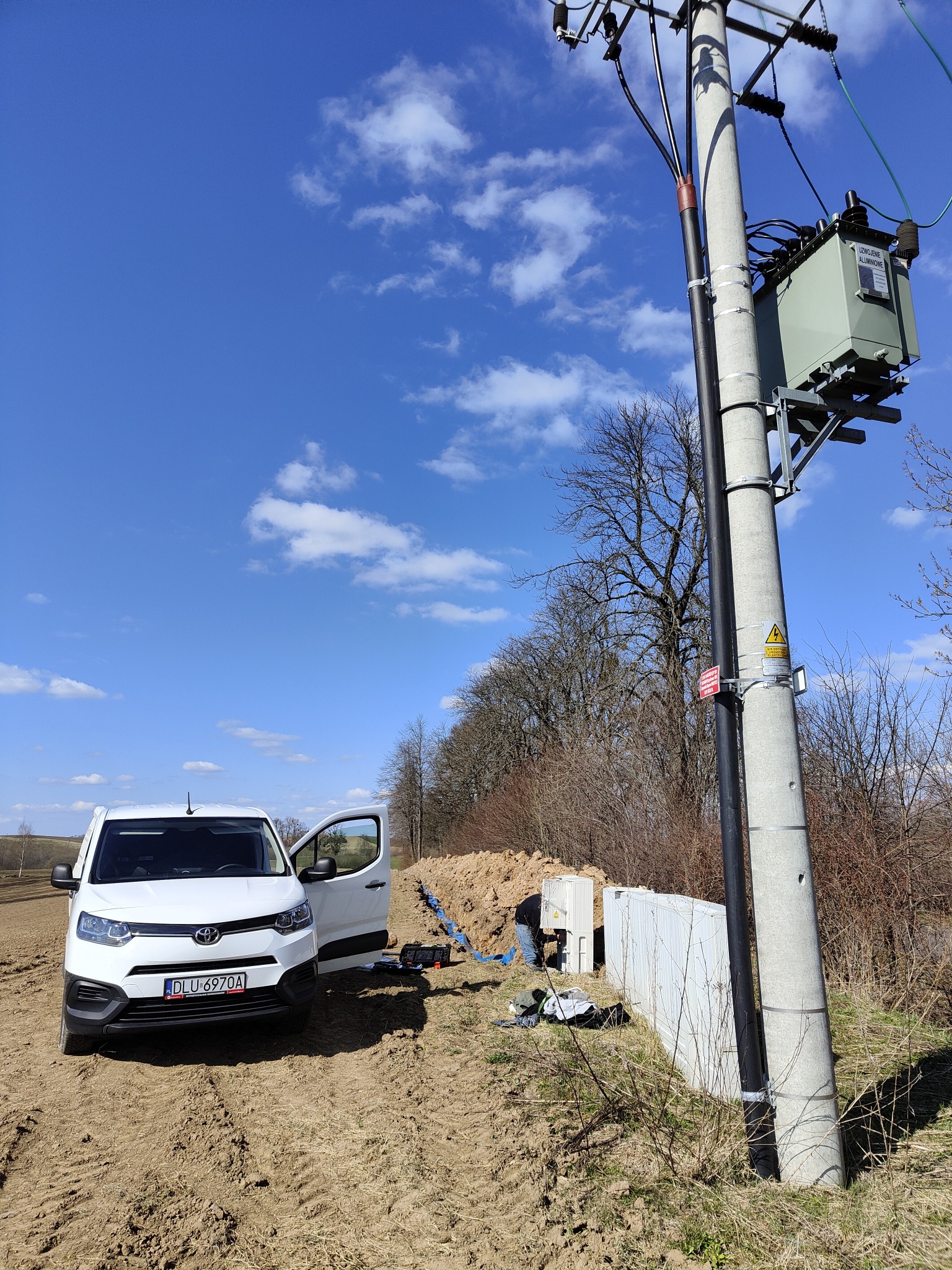 Biały samochód dostawczy Toyota obok betonowego słupa energetycznego z transformatorem, widoczne prace ziemne i instalacja kabli. Błękitne niebo z chmurami w tle.