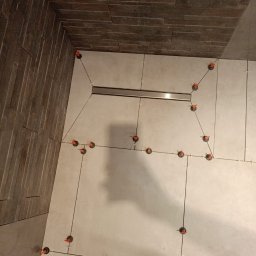 LuxBath Company LTD - Ułożone płytki podłogowe w łazience z widocznym odpływem liniowym i systemem poziomowania płytek. Ściana wykończona płytkami imitującymi kamień.