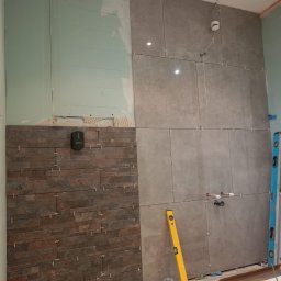 LuxBath Company LTD - Układanie płytek w łazience: szare płytki wielkoformatowe i dekoracyjne płytki imitujące kamień. Widoczny poziomica, laser krzyżowy i krzyżyki dystansowe.