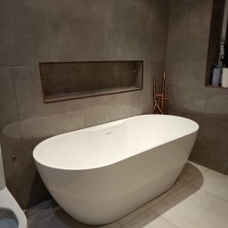 LuxBath Company LTD - Nowoczesna łazienka z wolnostojącą białą wanną, szarą płytką na ścianach i podłodze oraz miedzianą armaturą. W ścianie wnęka z półką.