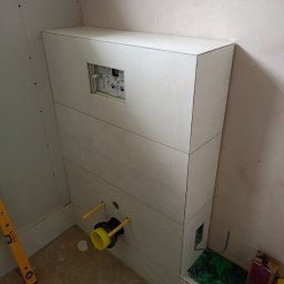LuxBath Company LTD - Obudowa stelaża WC z płyt gipsowych w trakcie montażu. Widoczne przyłącza i otwór rewizyjny. Poziomica na podłodze. Prace wykończeniowe w łazience.