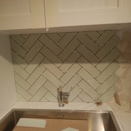 LuxBath Company LTD - Układanie jasnych płytek w jodełkę nad zlewem w kuchni. Widoczne płytki, fuga i armatura. Częściowo wykończona ściana nad blatem kuchennym.