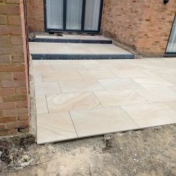 LuxBath Company LTD - Ułożona, beżowa, prostokątna kostka brukowa na tarasie przy ceglanym domu. Widoczne czarne obrzeża i fragmenty niezakończonych prac ziemnych.