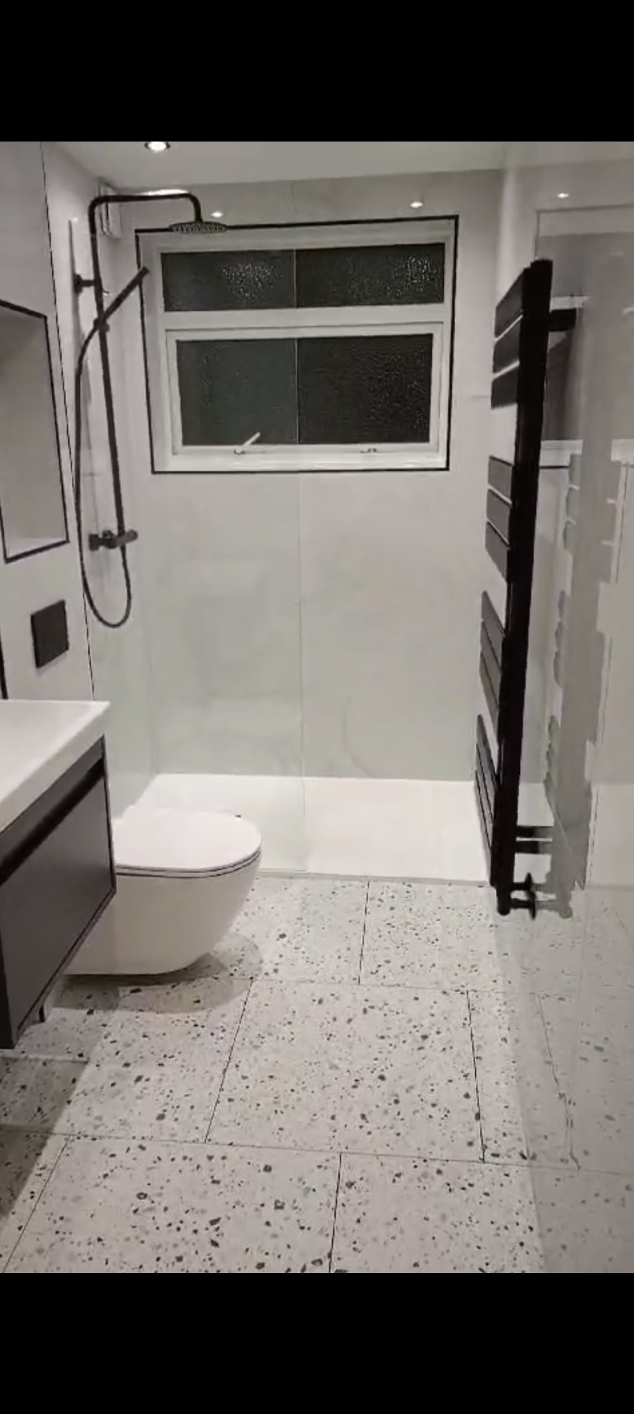 Nowoczesna łazienka z kabiną prysznicową typu walk-in, wiszącą toaletą i grzejnikiem drabinkowym w czarnym kolorze. Uwagę przyciągają płytki podłogowe w stylu lastryko.